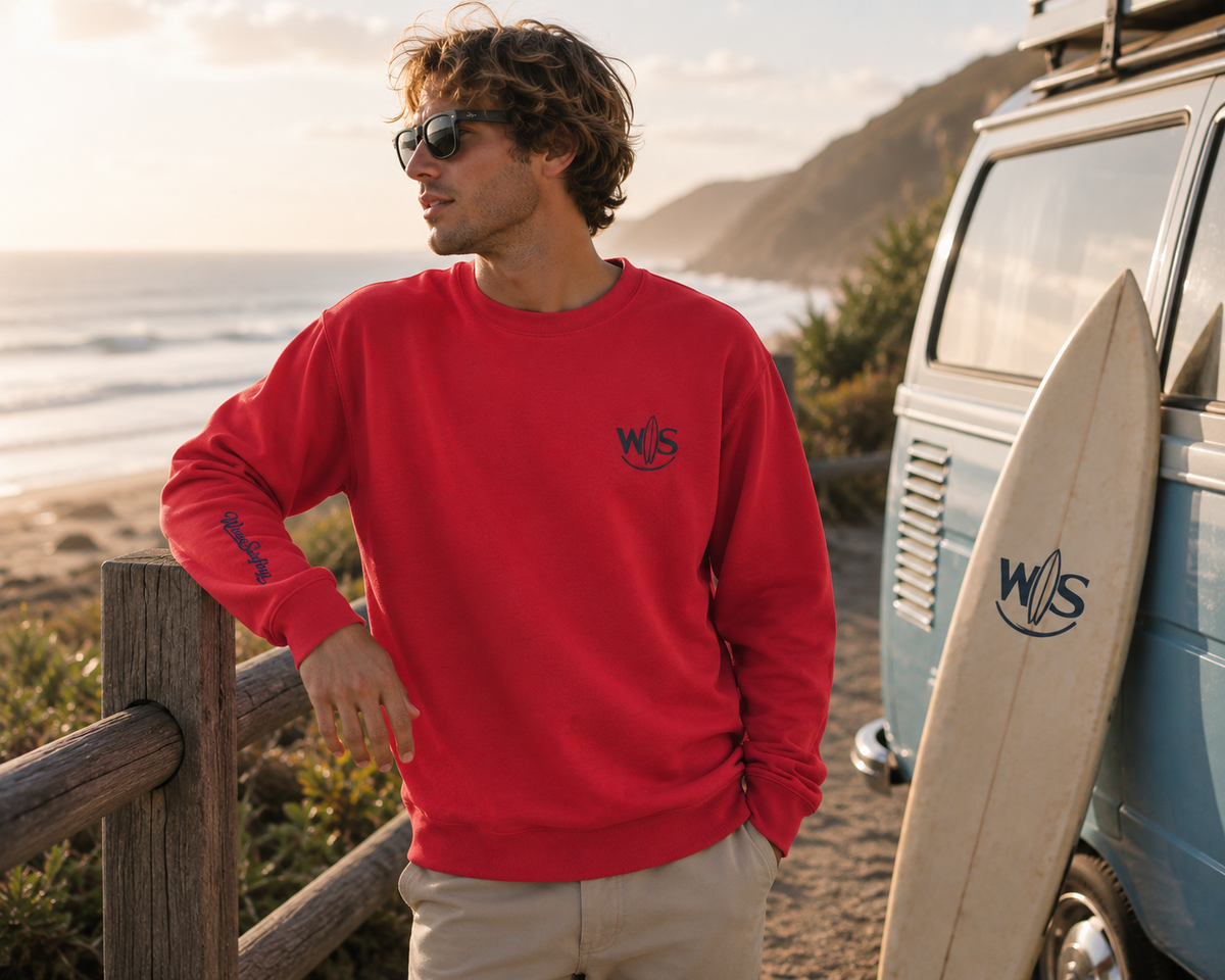 Man wearing a red WaveSurfing™ unisex premium sweatshirt with WS chest logo and WaveSurfing sleeve detail in a coastal beach setting at sunset.