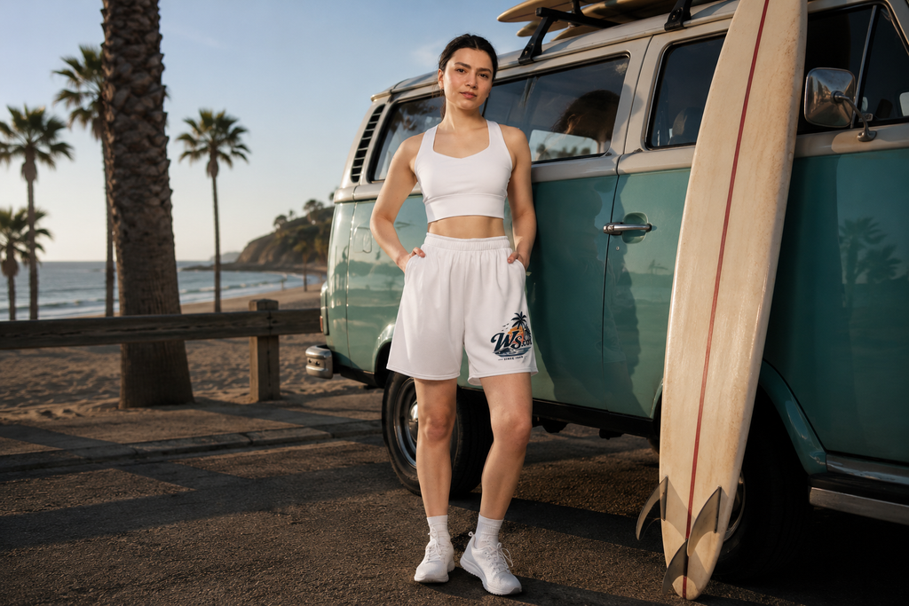 Woman wearing WaveSurfing™ white unisex mesh shorts standing beside a vintage surf van on a sunny beach. Lightweight athletic shorts with a relaxed fit, perfect for gym workouts, beach days, travel, and everyday coastal style.